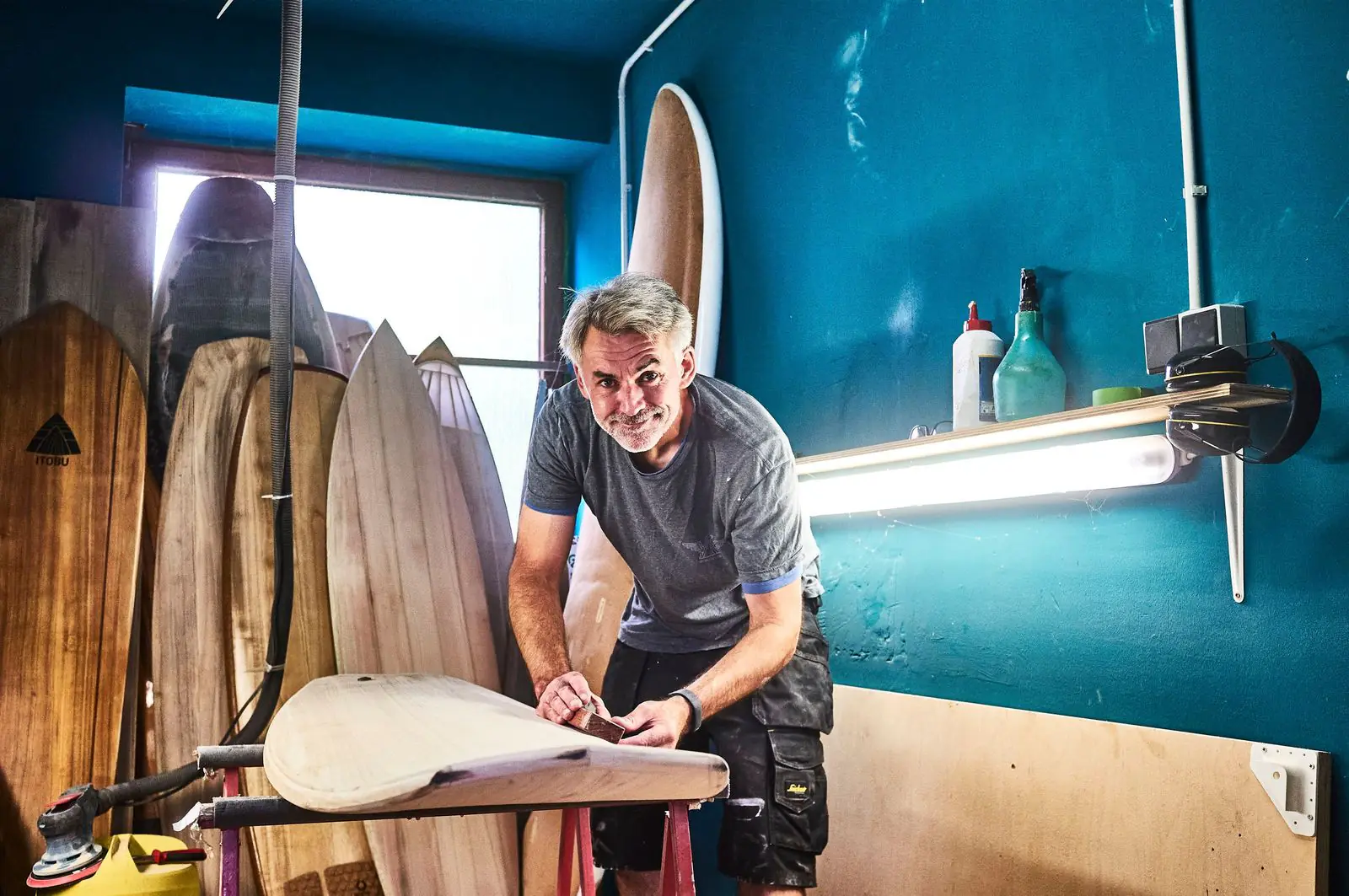 Tobias Herold im Shaperaum, er arbeitet an einem Surfbrett aus Holz, dahinter weitere Holzbretter an der türkisen Wand